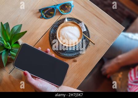 Hand einer Frau, die in einem Restaurant ein Mobiltelefon mit Kaffee und Sonnenbrille auf einem Holztisch hält. Möglichkeit, auf dem Bildschirm der Zelle phon setzen Stockfoto