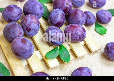 Frische rohe blaue und violette Pflaumen mit grünen Blättern auf hellem Holzgrund. Stockfoto