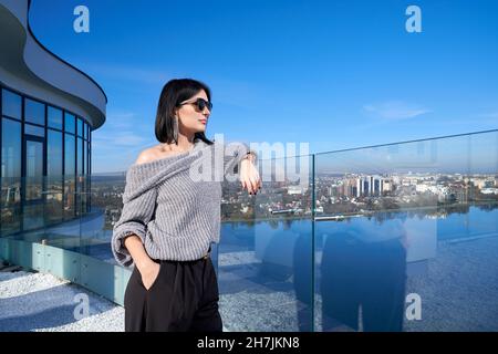 Atemberaubende Frau mit dunklem Haar, die auf der hohen offenen Terrasse steht und einen atemberaubenden Blick auf die große Stadt genießt. Kaukasische Frau trägt Sonnenbrillen und stilvolle Kleidung. Stockfoto