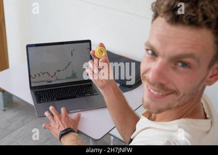 Der junge lächelnde Trader verdient Geld mit dem Kryptowährungshandel, hält eine Bitcoin-Münze und das Candlestick-Diagramm ist grün und positiv Stockfoto