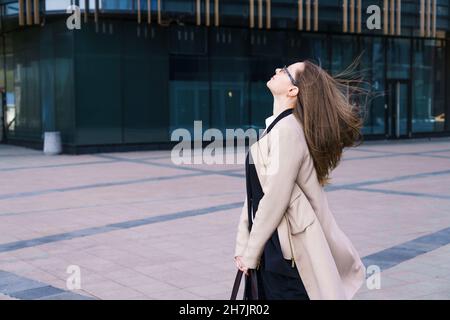 Eine Geschäftsfrau aus dem Kaukasus in einem Mantel und Anzug mit nachdenklicher Optik steht in einer Brille in der Nähe des Geschäftszentrums. Das Konzept der Lösung komplexer Geschäftsprobleme Stockfoto