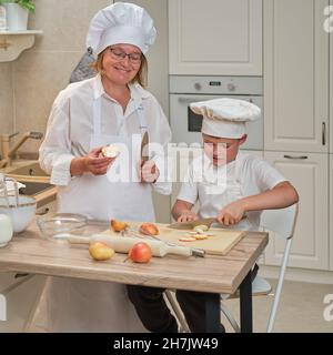 Mutter und Sohn in weißen Chefkleidern schnitten in ihrer eigenen Küche mit einem Messer Birnen. Eine Erwachsene Frau mit einem kleinen Jungen wird einen Kuchen kochen Stockfoto