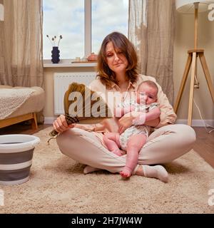 Eine traurige Frau mit einem Baby sitzt auf dem Boden in einem Wohnzimmer zu Hause, Probleme mit der Hausarbeit bei der Geburt eines Kindes Stockfoto