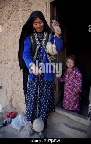 Eine ethnische Hazara-Frau, die mit einem Kind in der Nähe der Tür ihres Schlammhauses steht, wind Fäden aus reiner Wolle, um Pullover im Dorf Raksha zu weben Stockfoto