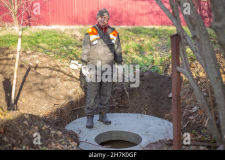 Bau einer Klärgrube für ein ländliches Haus. Ein Arbeiter in Overalls steht auf den Betonringen eines septischen Tanks mit einer Luke. Stockfoto