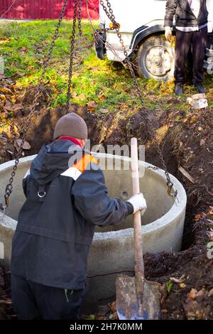 Bau einer Klärgrube für ein ländliches Haus. Ein Lader senkt Betonringe in eine Grube, um einen Klärgruben zu bauen. Der Arbeiter steuert die Installation. Stockfoto