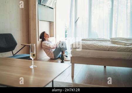 Traurige Frau, die auf dem Boden neben dem Fenster im Schlafzimmer mit einer geöffneten Flasche Alkohol saß. Unfokussiertes Weißweinglas auf dem Vordergrundtisch. Stockfoto