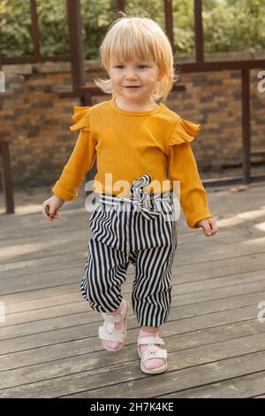 Ein schönes, fröhliches kleines Mädchen in einem stylischen gestreiften Anzug läuft in einem Stadtpark. Lifestyle Stockfoto