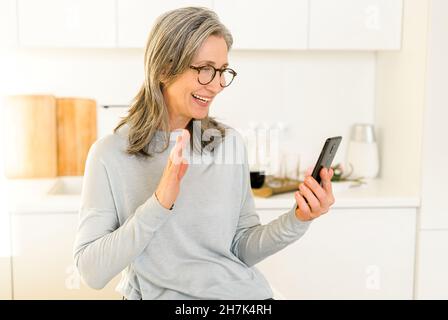 Glücklich lächelnde reife Frau, die in der Küche sitzt, sich entspannt und per Videoverbindung mit ihrem Freund oder ihrer Familie spricht, ältere grauhaarige Frau, die Mobiltelefon hält und die Hand winkt Stockfoto