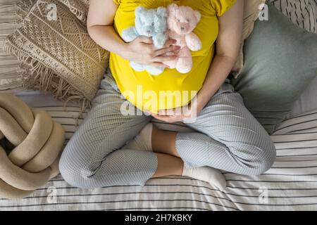 Draufsicht Schwangeren junge Frau hält gelb mit zwei niedlichen Bären Spielzeug auf dem Bauch sitzen auf dem Bett Stockfoto