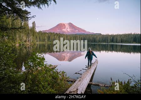 Frau, die auf einem Baumstamm mit Mount Adams im See reflektiert Stockfoto