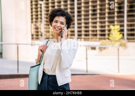 Glückliche junge, ethnische Managerin mit kurzen lockigen Haaren in elegantem Outfit, lächelnd und wegblickend, während sie auf dem Smartphone auf der Straße der Stadt spricht Stockfoto