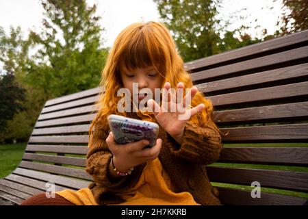 Rotschopf Mädchen mit Handy, während auf Parkbank sitzen Stockfoto