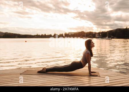 Frau, die während des Sonnenuntergangs am Steg am See Yoga praktiziert Stockfoto