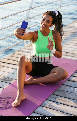 Lächelnde junge Sportlerin, die beim Selfie über das Smartphone das Friedenszeichen gestikulierte Stockfoto