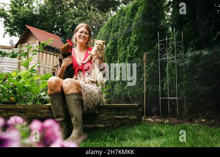 Glückliche Frau, die Katze und Hahn trägt, während sie im Hinterhof sitzt Stockfoto