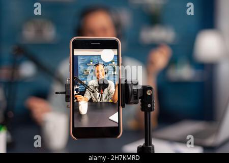 Nahaufnahme der Handy-Aufnahme lächelnde Frau vlogger auf Live-Stream Podcast. Influencer mit Mikrofon und Kopfhörern, im Gespräch vor der Telefonkamera und Filmen von Vlog für Social Media-Kanal. Stockfoto