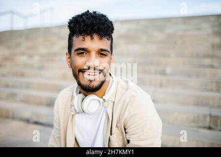 Lächelnder junger Mann mit schwarzen Locken und kabellosen Kopfhörern Stockfoto