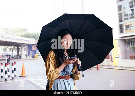 Junge Frau mit Regenschirm und Smartphone auf der Straße Stockfoto