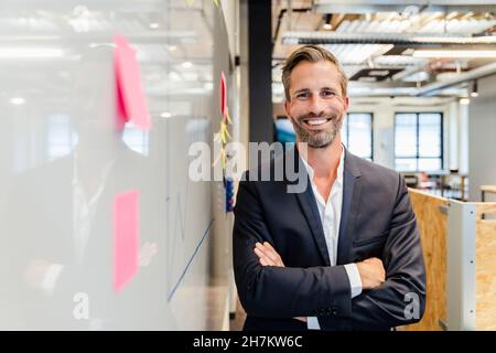 Lächelnder bärtiger Geschäftsmann mit gekreuzten Armen im Büro Stockfoto