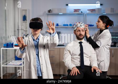 Neurologe Arzt trägt Virtual-Reality-Headset, während Forscherin Frau eeg-Scanner eines Mannes Patienten anpassen Gehirnaktivität während Neurologie Experiment analysieren. Wissenschaftler Ingenieur mit High-Tech Stockfoto