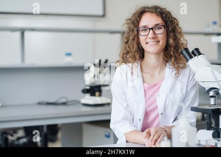 Lächelnder junger Wissenschaftler mit Mikroskop am Labortisch Stockfoto