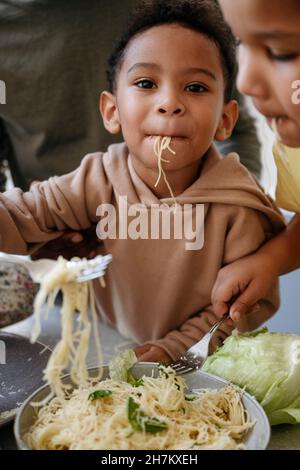 Netter Junge, der Spaghetti zwischen Bruder und Vater in der Küche isst Stockfoto