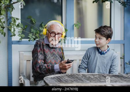 Lächelnder Junge, der Großvater beim Musikhören über Kopfhörer im Hinterhof ansieht Stockfoto