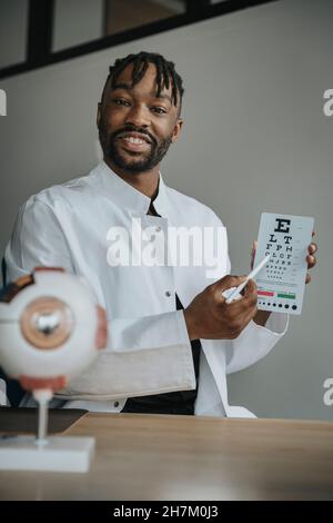 Der junge Augenarzt hält ein Alphabetdiagramm am Schreibtisch Stockfoto
