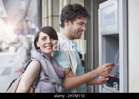 Lächelnder Mann, der Geld vom Geldautomaten abzieht, der neben der Freundin steht Stockfoto