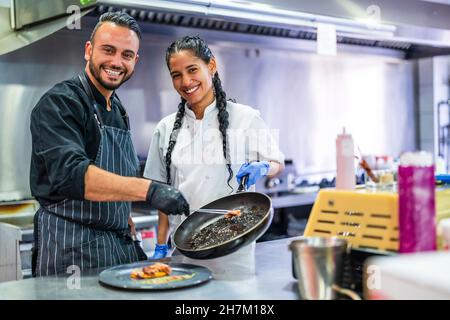 Lächelnder Küchenassistent hilft dem Koch bei der Zubereitung von Speisen im Restaurant Stockfoto