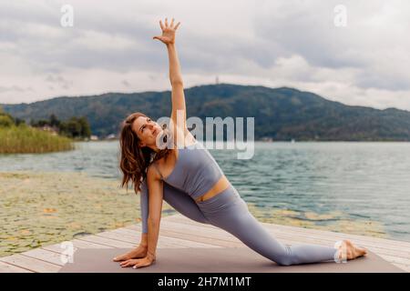 Frau mit erhobener Hand praktiziert Yoga vor dem See Stockfoto