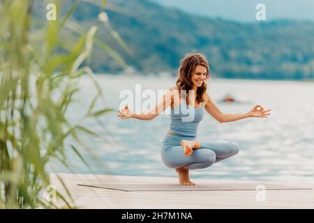 Glückliche Frau, die Yoga auf dem Steg am See macht Stockfoto