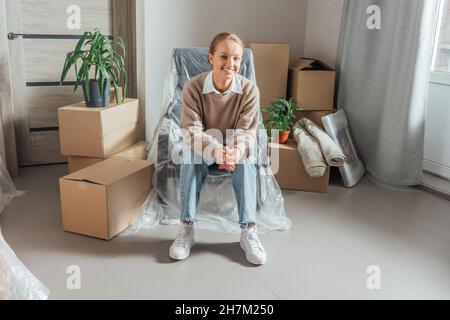 Lächelnde junge Frau, die zu Hause auf einem in Plastik gewickelten Sessel sitzt Stockfoto