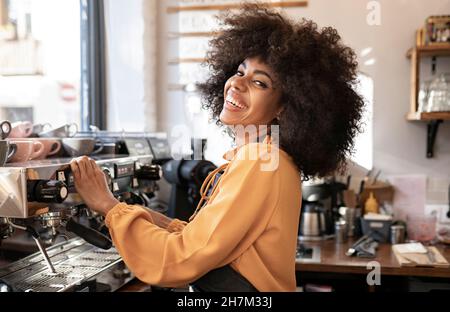 Lächelnde Kellnerin mit afro Frisur, die an der Kaffeemaschine steht Stockfoto