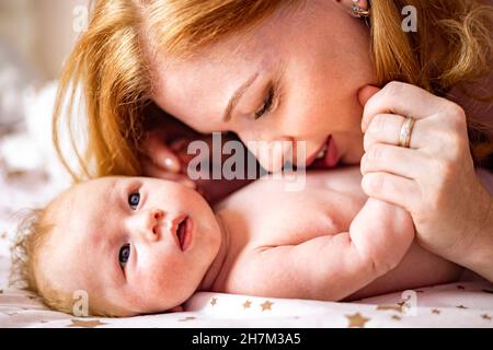 Glückliche Rothaarige Mutter umarmt kleines neugeborenes Baby, das zusammen auf Baumwollbett liegt und Mutterschaft genießt Stockfoto