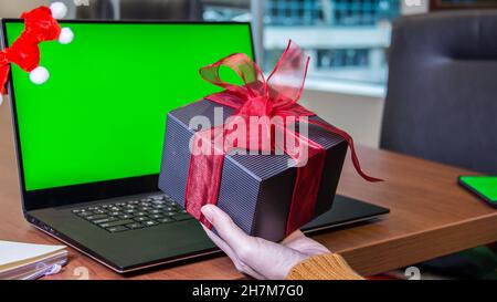 Hand hält große Geschenkbox neben Laptop mit grünem Bildschirm mockup, weihnachtseinkäufe. Online-Shopping. Neujahr. Feier, Arbeit von Home offic Stockfoto