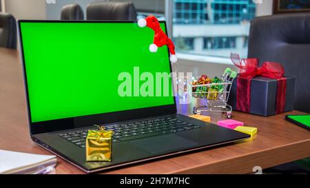 Laptop mit grünem Bildschirm Mockup, Warenkorb voller Geschenke auf dem Hintergrund. Online-Shopping Stockfoto