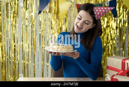 Eine fröhliche junge Brünette in einem festlichen Hut hält mit einer brennenden Kerze einen Kuchen in den Händen und genießt den Urlaub. Geburtstagskonzept. Stockfoto
