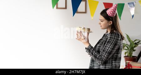 Eine junge kaukasische Frau steht in einem weiblichen Anzug, hält einen Kuchen in ihren Händen und schaut ihn an. Geburtstagskonzept im Büro. Baner. Stockfoto