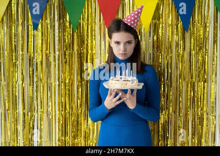 Eine traurige Frau auf gelbem Hintergrund in einem Partyhut steht und hält einen Kuchen in den Händen. Geburtstag langweilige Party-Konzept. Stockfoto