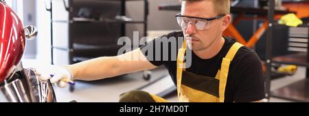 Mann, der in der Autowerkstatt Farbe an das Motorrad abholt, in einer Brille Stockfoto