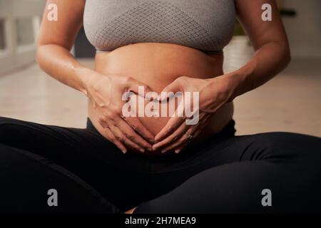 Die kaukasische Schwangerenmutter sitzt auf einer Yogamatte und formt eine Herzform mit den Händen auf dem Bauch Stockfoto