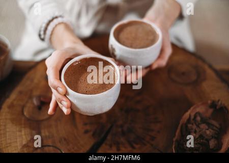 Heißer handgemachter zeremonieller Kakao in weißen Tassen. Frau Hände geben Handwerk Kakao, Draufsicht auf Holztisch. Bio gesundes Schokoladengetränk aus zubereitet Stockfoto