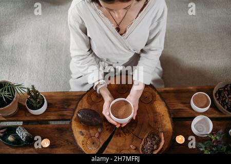 Heißer handgemachter zeremonieller Kakao in weißer Tasse. Frau Hände halten Handwerk Kakao, Draufsicht auf Holztisch. Bio gesundes Schokoladengetränk aus zubereitet Stockfoto