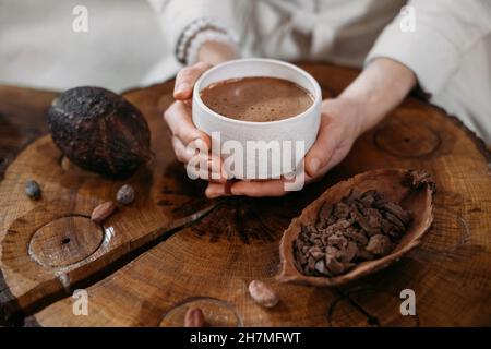 Heißer handgemachter zeremonieller Kakao in weißer Tasse. Frau Hände halten Handwerk Kakao, Draufsicht auf Holztisch. Bio gesundes Schokoladengetränk aus zubereitet Stockfoto