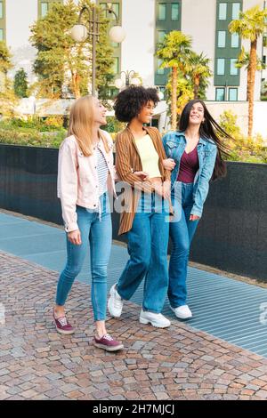 Ein ganzer Körper junger multirassischer Freundinnen in Jeans, die sich fröhlich unterhalten und lachen, während sie auf einem gepflasterten Gehweg in der Sommerstadt zusammen spazieren Stockfoto