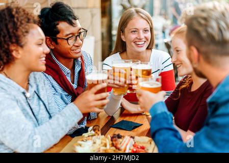 Eine Gruppe verschiedener junger Freunde, die gemeinsam ein Bier trinken und dabei ihre Gläser mit Fokus auf eine lächelnde junge blonde Frau anstoßen Stockfoto