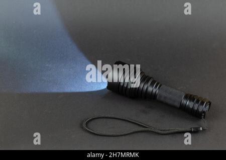 Hochleistungs-LED-Handheld-Taschenlampe beleuchtet den Raum vor Ihnen. Kleine, moderne taktische Handtaschenlampe mit einem Gurt gegen ein Grau Stockfoto