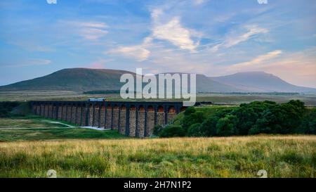 Landschaftlich reizvolle Tal- und Sonnenuntergänge (Lokomotive, Wahrzeichen Ribblehead Viaduct Torbögen, hohe Hügel und Berge) - North Yorkshire Dales, England. Stockfoto